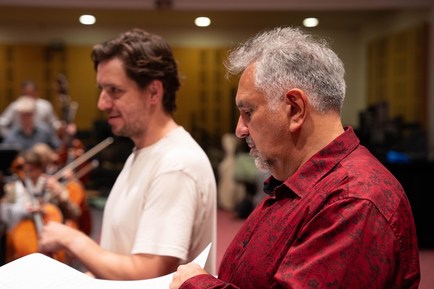 A grey-haired man sits next to a dark-haired man near an orchestra in a rehearsal space.