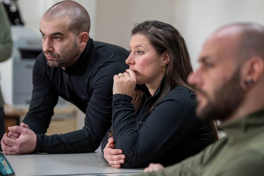 A woman and two men lean over a table listening intently 