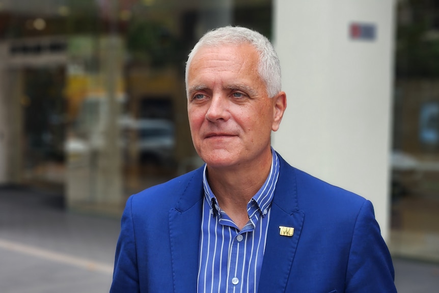 A middle-aged man with short grey hair wears a suit while standing on a city street.