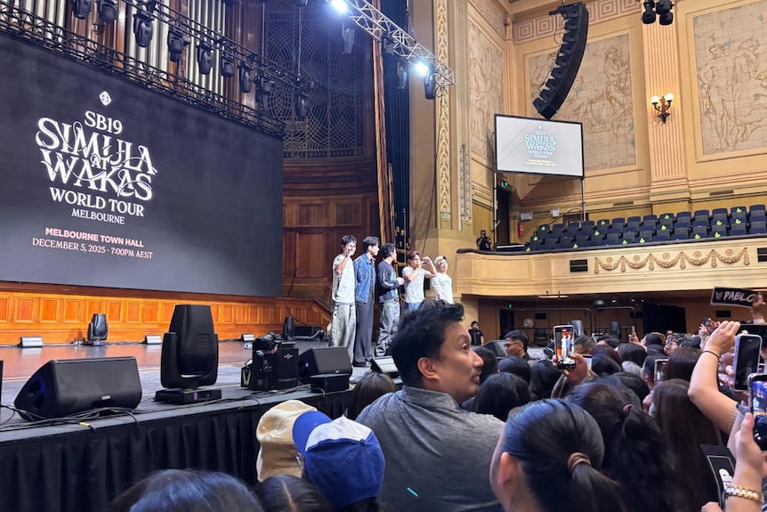 A group of boys stand on stage in front of a large screen with the name of their tour.