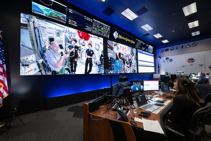 A large wall of screens with astronauts in a big room with computer-covered desks and headset-wearing NASA staff.