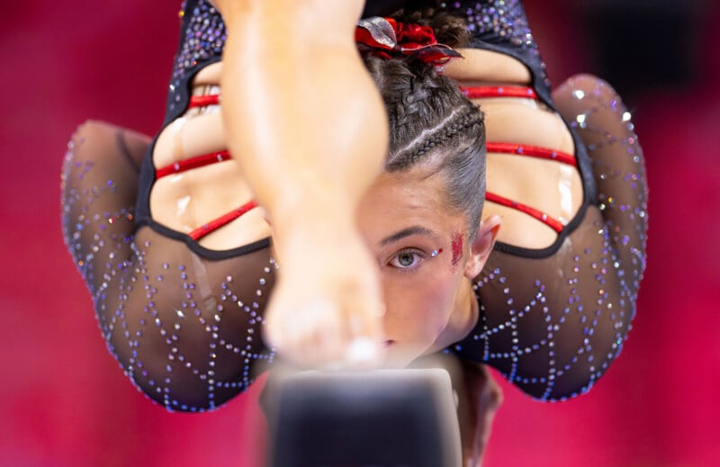 A gymnast with braided hair and a sparkly black leotard performs an intense balancing pose on a beam, her face focused and partially framed by her leg, against a blurred red background.
