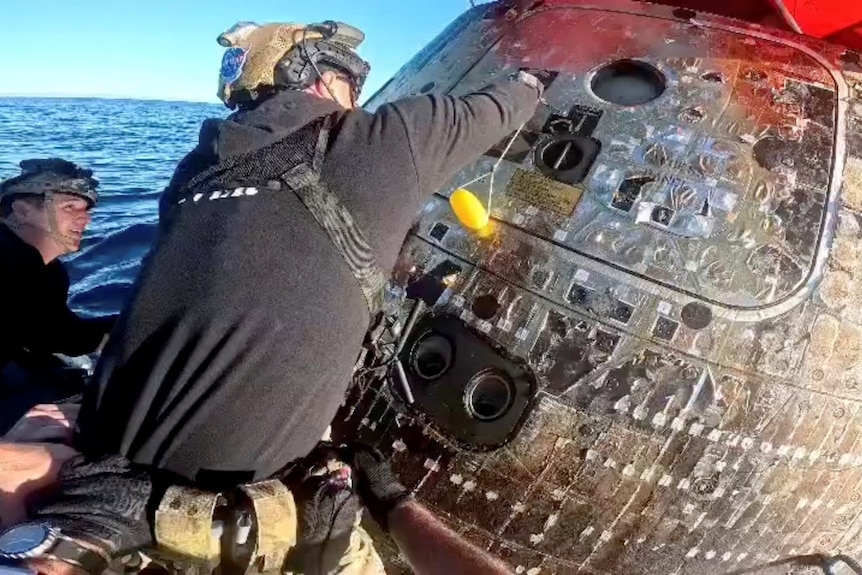 Two members of the Navy recovery team, wearing protective helmets, are about to open the hatch to a space capsule.