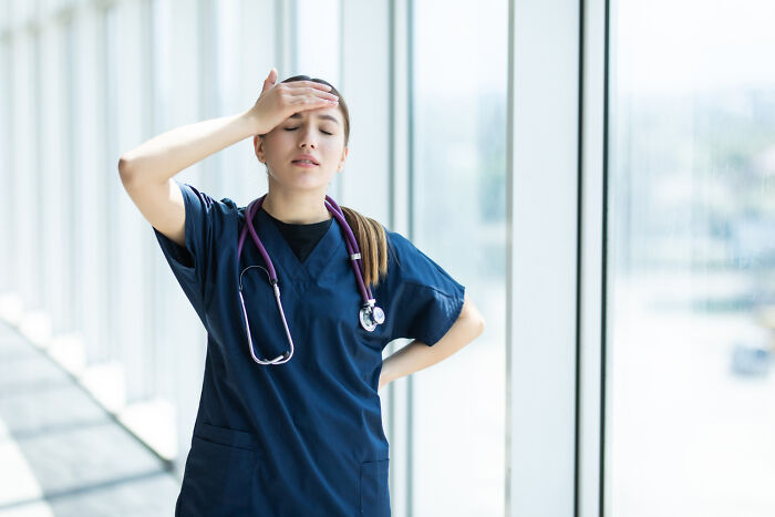 Female nurse in blue scrubs with stethoscope, looking stressed and overwhelmed inside a bright hospital corridor.