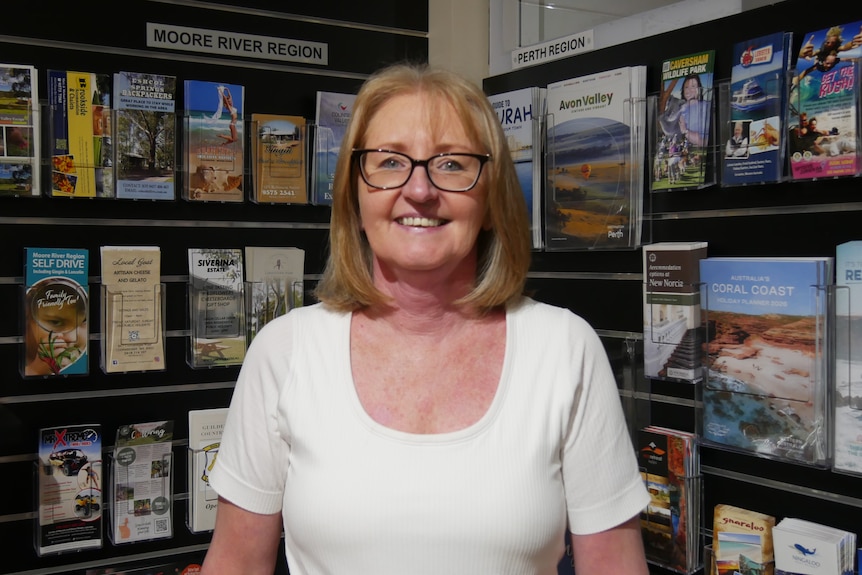 A woman in glasses stand infront of tourism brouchures smiling at the camera.