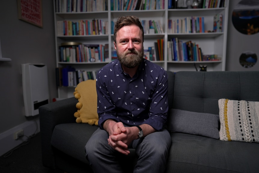 A man with a beard wearing a business shirt and trousers sits on a grey couch in front of a bookcase.