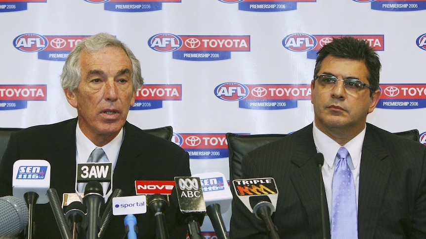 Two senior AFL executives sit in front of a bank of microphones, explaining an AFL Commission decision to the media.