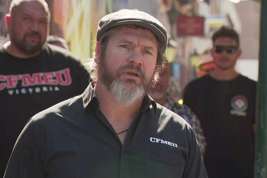 A man in a cap and grey-brown beard speaks at a media conference. A man in a CFMEU t-shirt stands behind him.