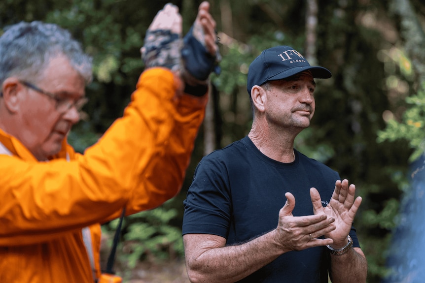 A man wearing a black T-shirt and cap clapping. Foreground a man in orange long-sleeve shirt also claps above his head