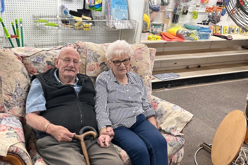 Mr and Mrs Pickles holding hands and sitting on couch inside their hardware store.