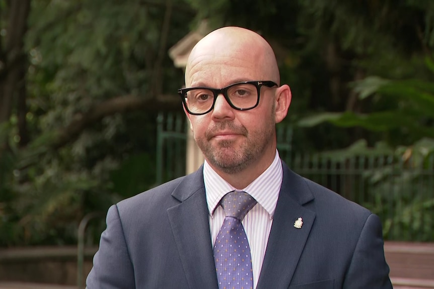 A man in a suit and thick rimmed glasses standing in front of Botanic Gardens.