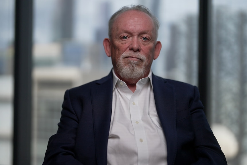 An older man with a short grey beard wearing a white business shirt and navy suit jacket sits in a chair in a high-rise office.