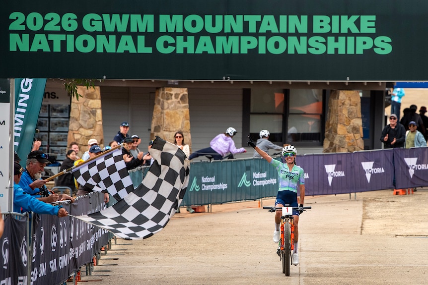 Bec Henderson crosses the line next to a chequered flag and waves her hand