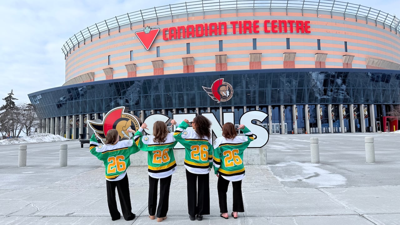Four girls stand with their backs towards the camera in front of the Canadian Tire Centre, an arena in Ottawa