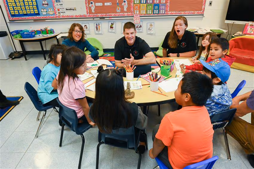 3 adults sit with children at school table 