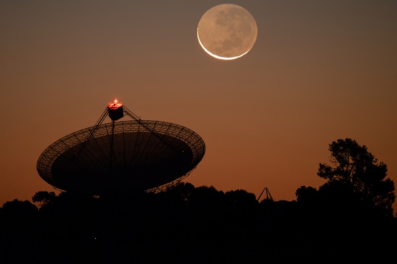 Murriyang, CSIRO’s Parkes radio telescope, in silhouette in the foreground under a dusky orange sky with a crescent Moon above. 