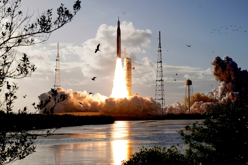 A long thin rocket, lifgts off vertically from a platform. Bright orange flames are left behind and huge clouds of smoke. 