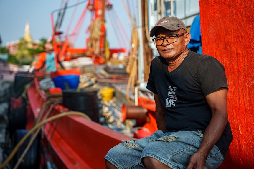 A man in a black t-shirt and jeans sits at the edge of a fishing boat.