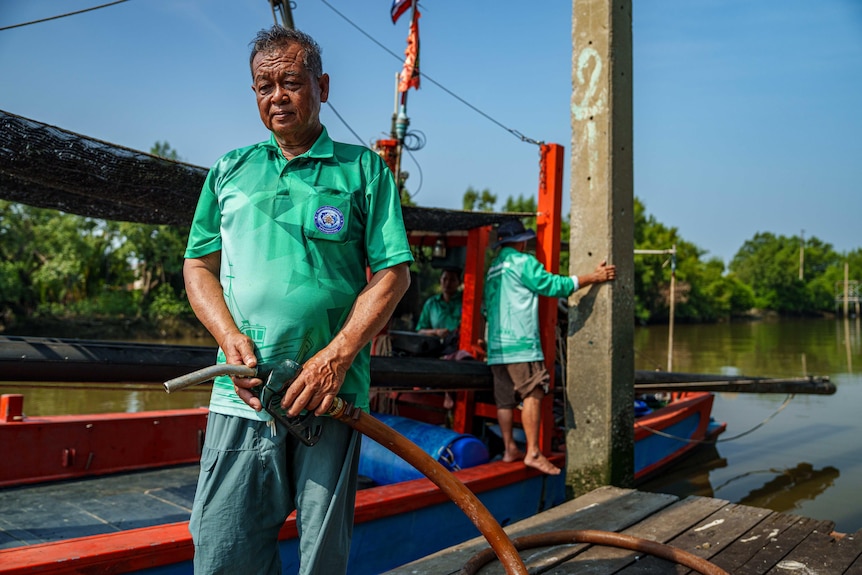 A man in a green shirt stands on a dock, looking solemn, holding the nozzle of a petrol pump.