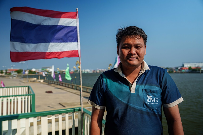 A man in a blue shirt stands at a jetty, with a Thai flag blowing in the wind beside him.