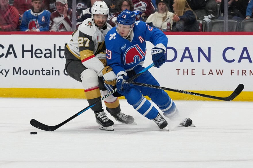Two hockey players, one in a white jersey, the other wearing blue, skate after a puck.