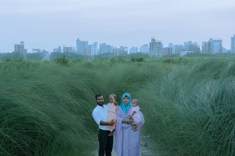 A family of four stands in tall green grass with city buildings in the background. The father holds a young child, while the mother, wearing a blue hijab, holds another child. The sky is overcast.