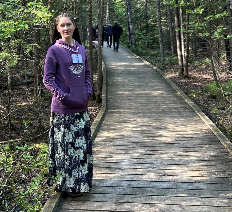 A teenaged girl wearing a long skirt and purple sweatshirt stands on a wooden boardwalk, surrounded by forest