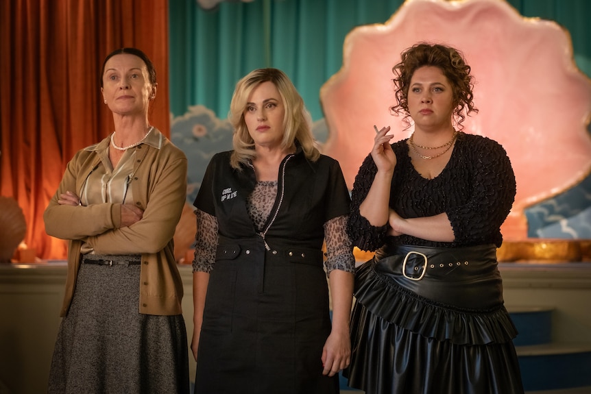 Three women stand in front of a stage with blue and red velvet curtains