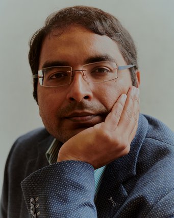 A portrait of Krishna Gummadi wearing rimless square glasses and a blue jacket, his chin resting on his hand.