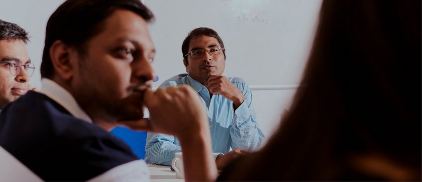 Krishna Gummadi, wearing rimless square glasses and a blue-and-white striped shirt, at a team meeting. In the foreground, three other colleagues are visible in the background or in half-profile, appearing blurry and partially in shadow.