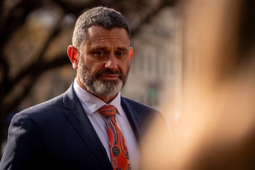 A man wearing a suit and tie with a First Nations pattern on it looks off camera with a serious expression