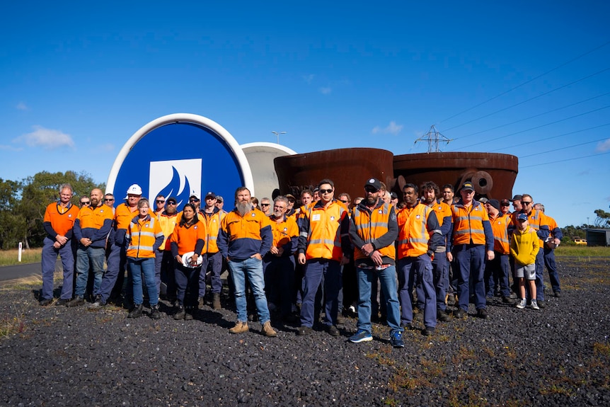 men and women in orange high vis outside a blue logo and a rusted sculpture