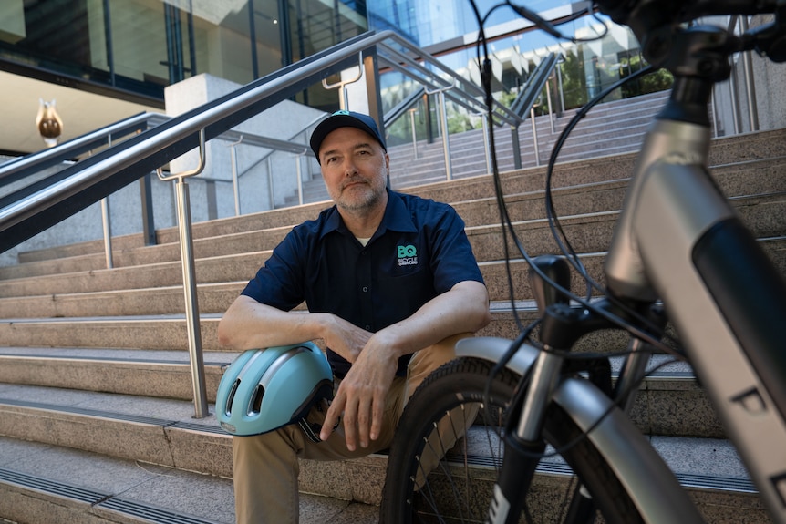 A man sits on outdoor steps with a bike helmet on his knee and an e-bike next to him