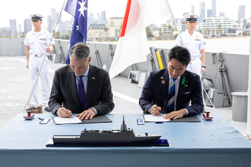 Richard Marles and Koizumi Shinjiro sit side by side onboard a warship signing documents.