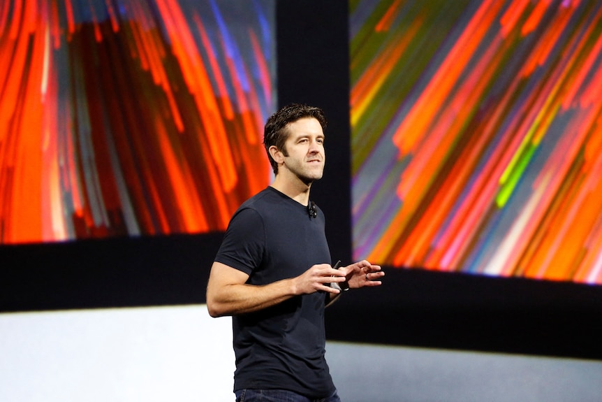 John Ternus gestures with his hands, wears dark tee, short dark hair, Caucasian, large screen behind with colours.