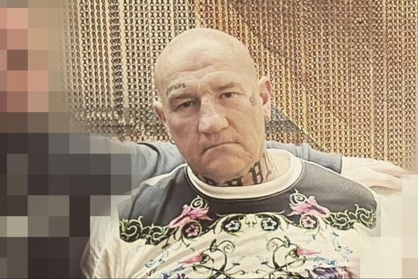 A pale photo of an unsmiling, bald man between two blurred figures.