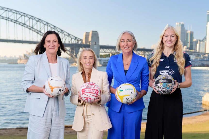 Four women stand holding netballs in front of Sydney Harbour