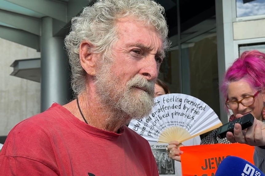 A man with grey hair and beard in a red shirt standing outside court