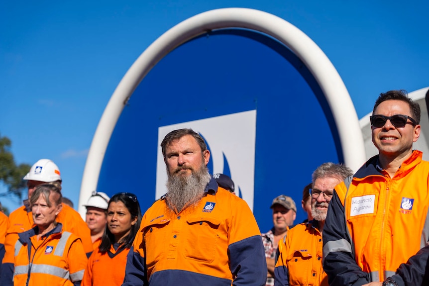 men and women in orange high vis outside a blue logo and a rusted sculpture