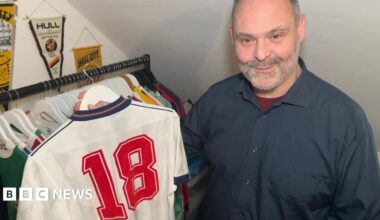 An man with cropped grey hair and stubble holds an original white England shirt from 1988, with the number 18 on the back, on a hanger. There is a rail of shirts behind him and Hull City pennants hanging on the wall. He is wearing a navy coloured short with a burgundy undershirt