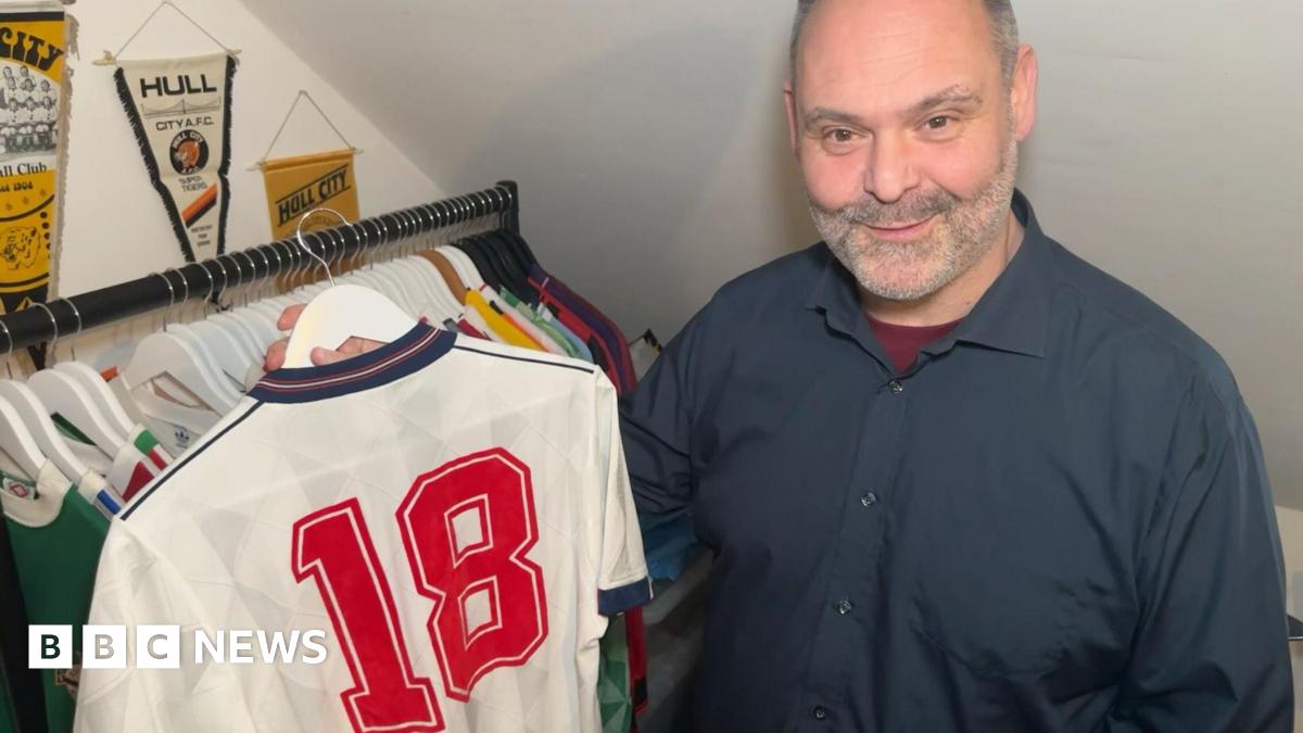 An man with cropped grey hair and stubble holds an original white England shirt from 1988, with the number 18 on the back, on a hanger. There is a rail of shirts behind him and Hull City pennants hanging on the wall. He is wearing a navy coloured short with a burgundy undershirt