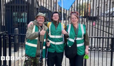 Rob, Lisa and son Drew outside gates in Downing Street. They are wearing dark green jackets and Rob Collins on the left is putting his thumb up.