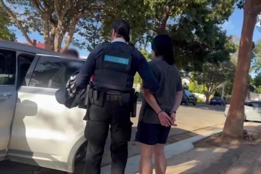 A man with hands cuffed behind him being escorted by a police officer into the backseat of a car parked on a suburban street