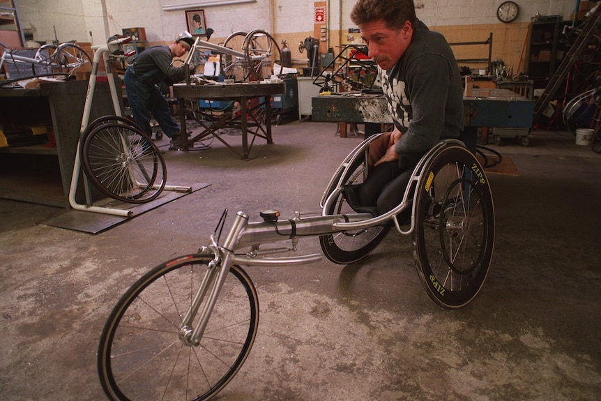 A photo of a younger Bob Hall in workshop, sitting a three-wheeled chair designed for racing.