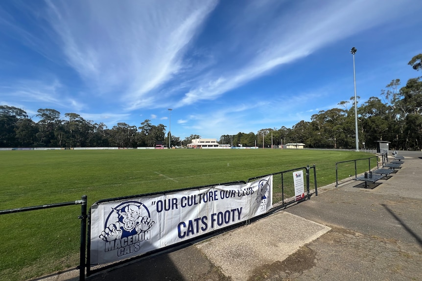 A green oval with shadows of trees in the foreground and blue sky and banner on the fence.