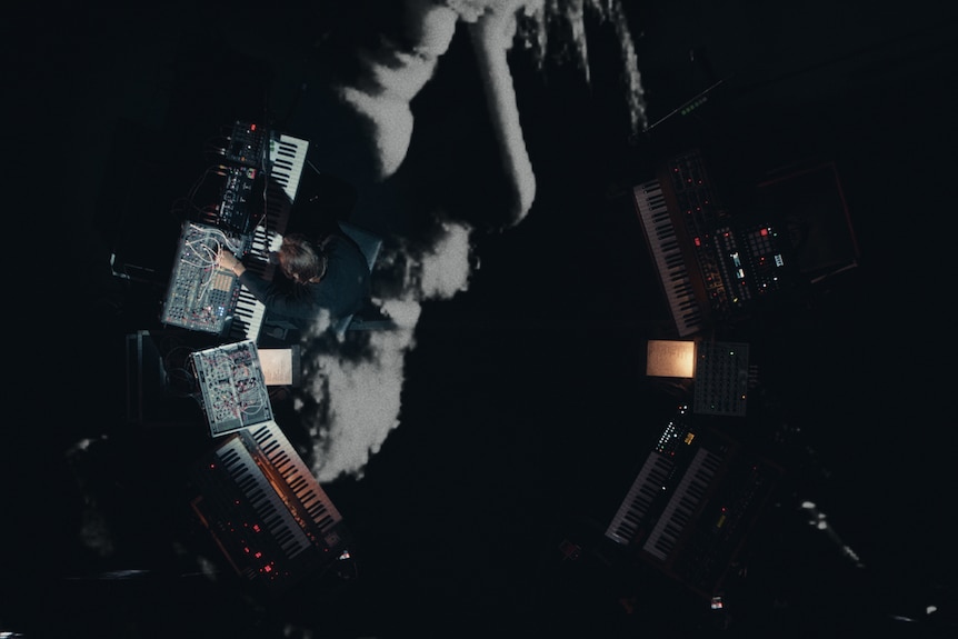 Outline of a bearded man with eyes closed overlaid with overhead shot of same man playing keys surrounded by synths and gear