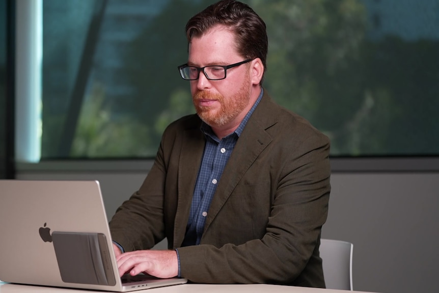 A dark-haired man in a dark blazer types on a laptop while sitting at  a desk in an office.