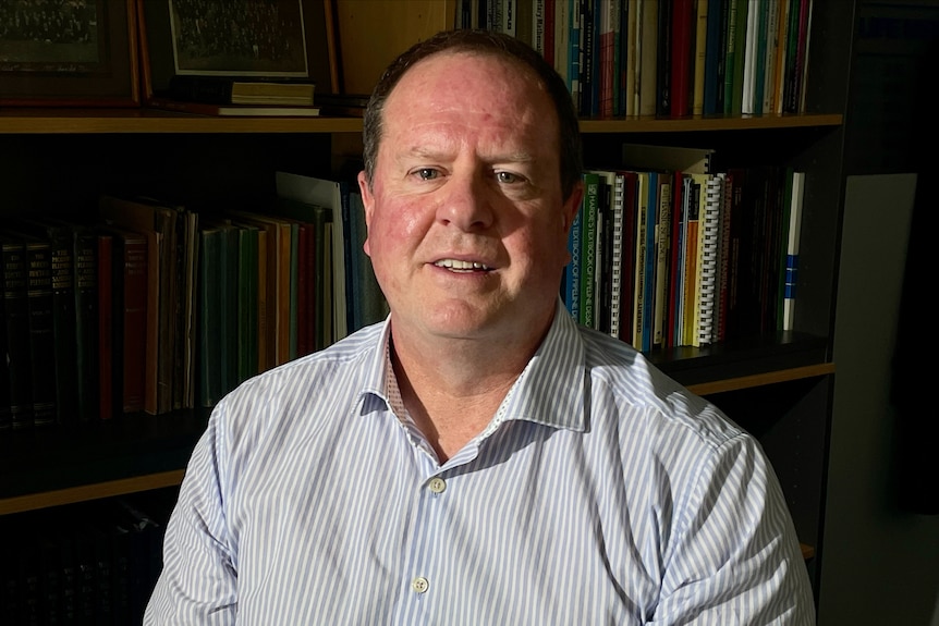 A middle-aged, dark-haired man sits in front of a set of bookshelves.
