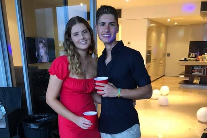 A smiling young blonde woman in a brightly-coloured dress cuddles up with a smiling young dark-haired man for in a big house.