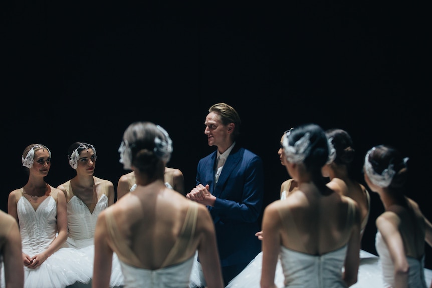 David Hallberg stands in a suit surrounded by ballerinas in white tutus